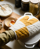 mustard yellow and white quilted oven mitt opening white dutch oven lid with wooden cutting board nearby