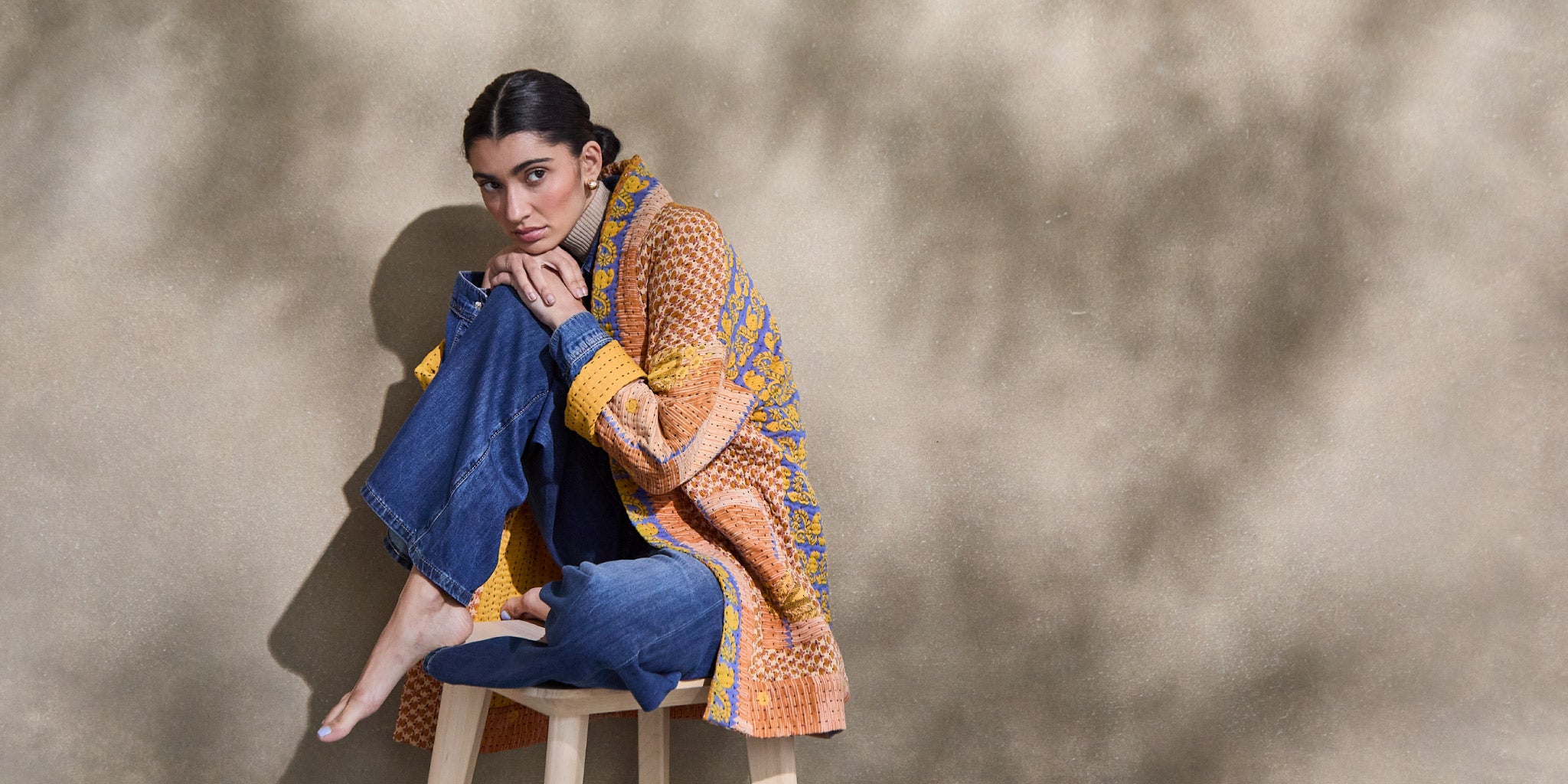 woman wearing kantha quilted jacket in orange and blue tones sitting on wooden stool