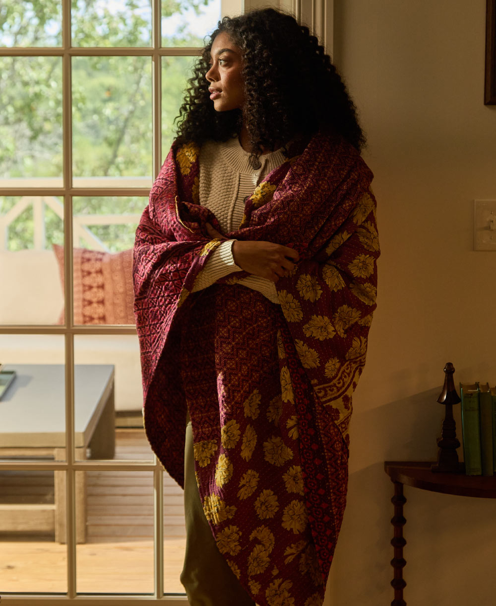 woman wrapped in red kantha quilt standing by window