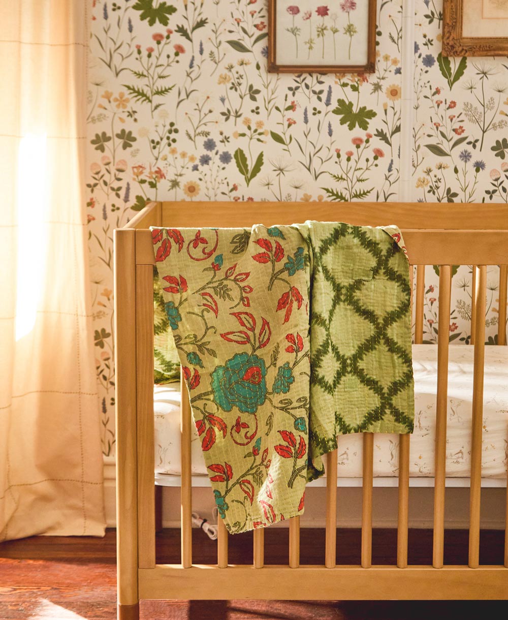 green floral small kantha quilt draped over light wooden modern baby crib with white floral wallpaper in background