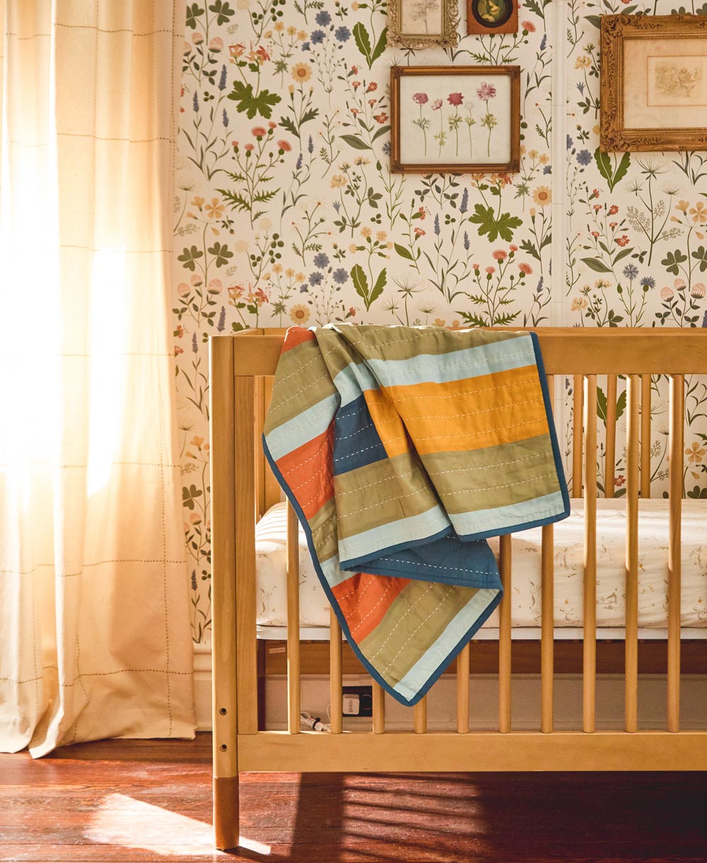 rainbow striped baby quilt draped over natural wood modern crib in nursery with floral wallpaper