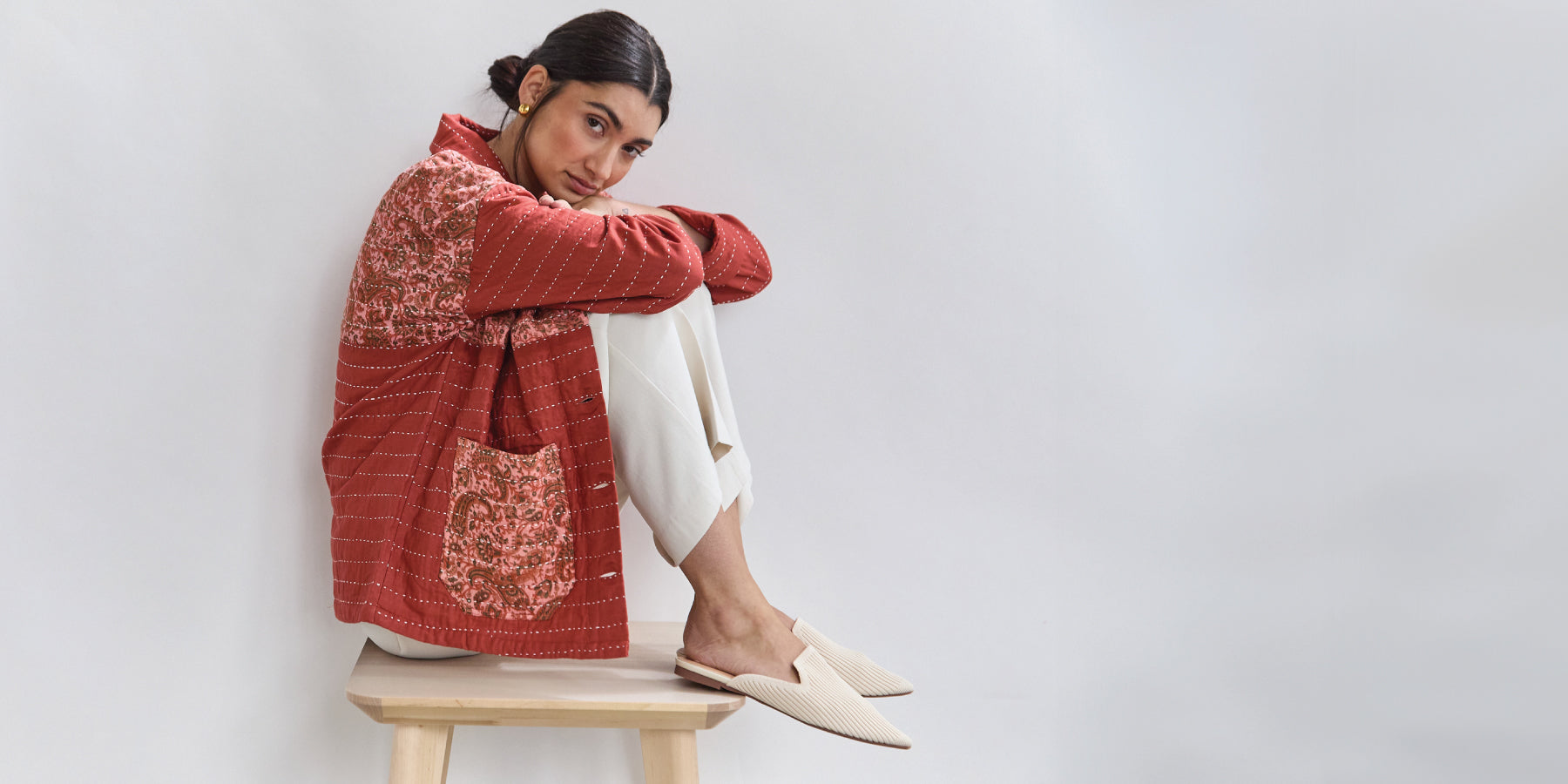 Woman wearing a red patterned jacket and white pants sitting on a stool against a light gray background