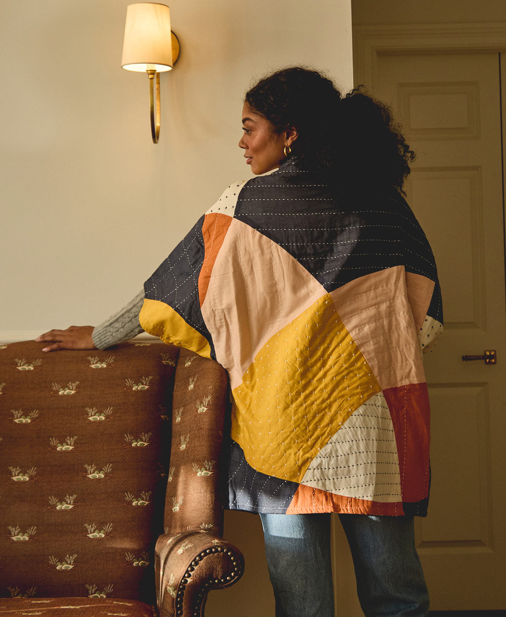 woman wearing abstract colorful cocoon quilted jacket standing next to a brown upholstered chair