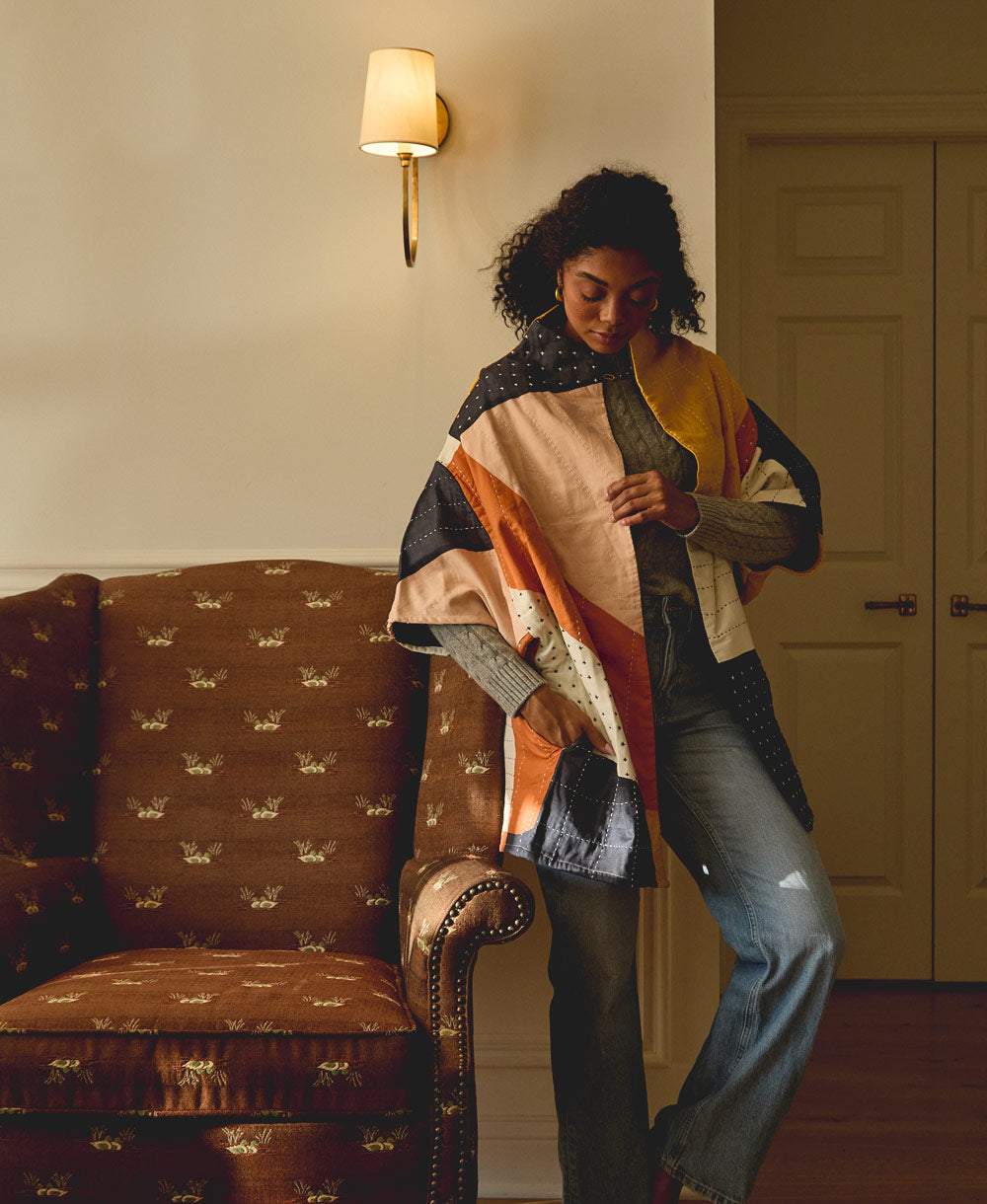 woman wearing colorful quilted jacket with hand-stitched embroidery standing next to vintage brown upholstered chair