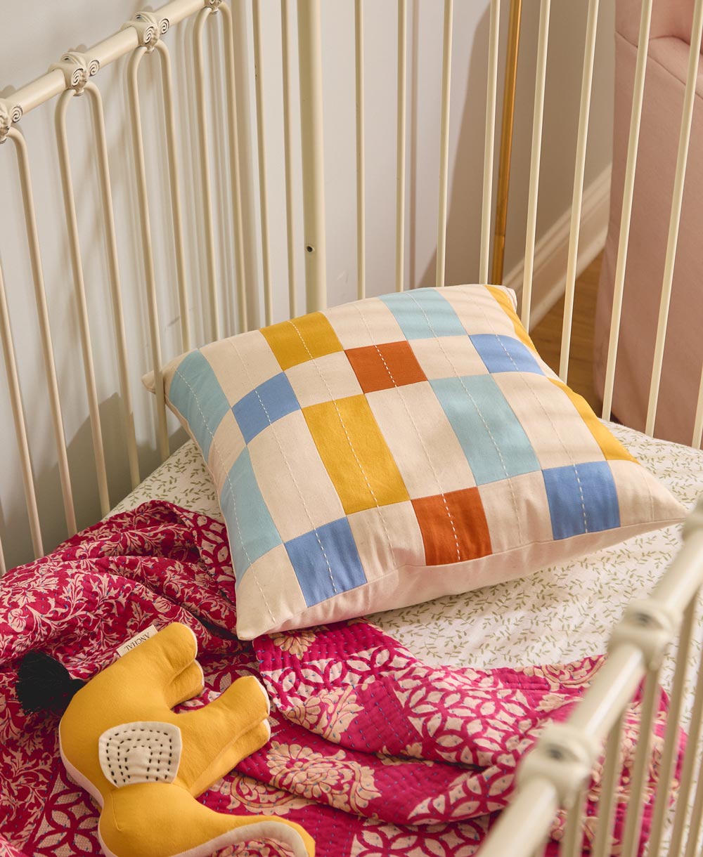 Colorful checkered pillow on a crib with a pink blanket and yellow stuffed elephant