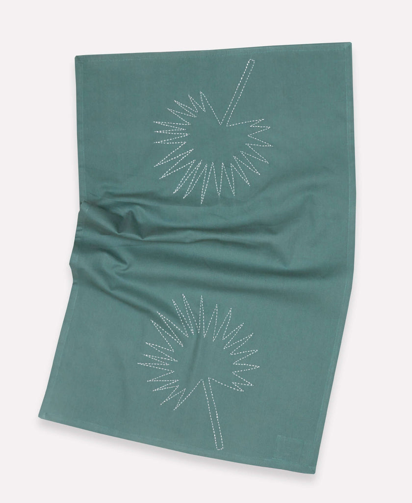 Flat-lay image of a green/blue tea towel with a leaf pattern in white stitching  
