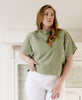 woman in sage green hand stitched crop top and white jeans standing in front of a white wall and fireplace