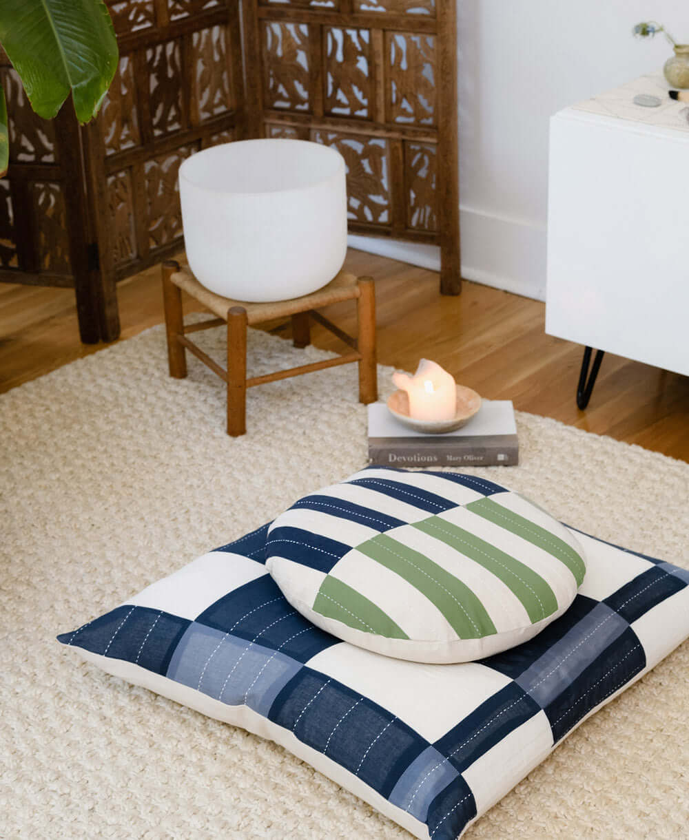 Circle throw pillow with navy and green stripes on top of larger square pillow with navy blue square and rectangle design on floor on white mat next to book with orange crystal on it