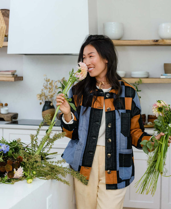 Woman holding and smelling flower wearing chore jacket made from organic cotton featuring blue, orange, and black plaid design