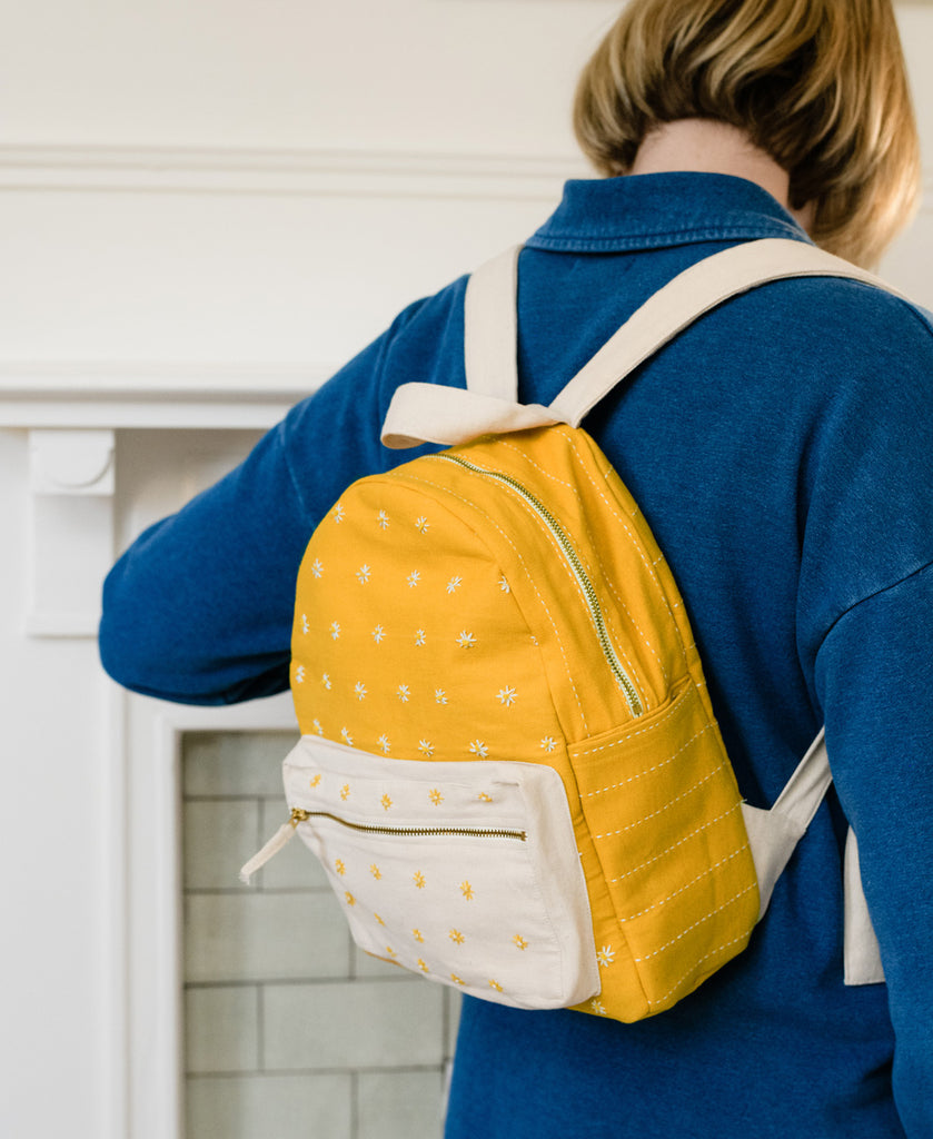 woman in blue shirt wears the mustard yellow and white small backpack with delicate hand embroidered daisies